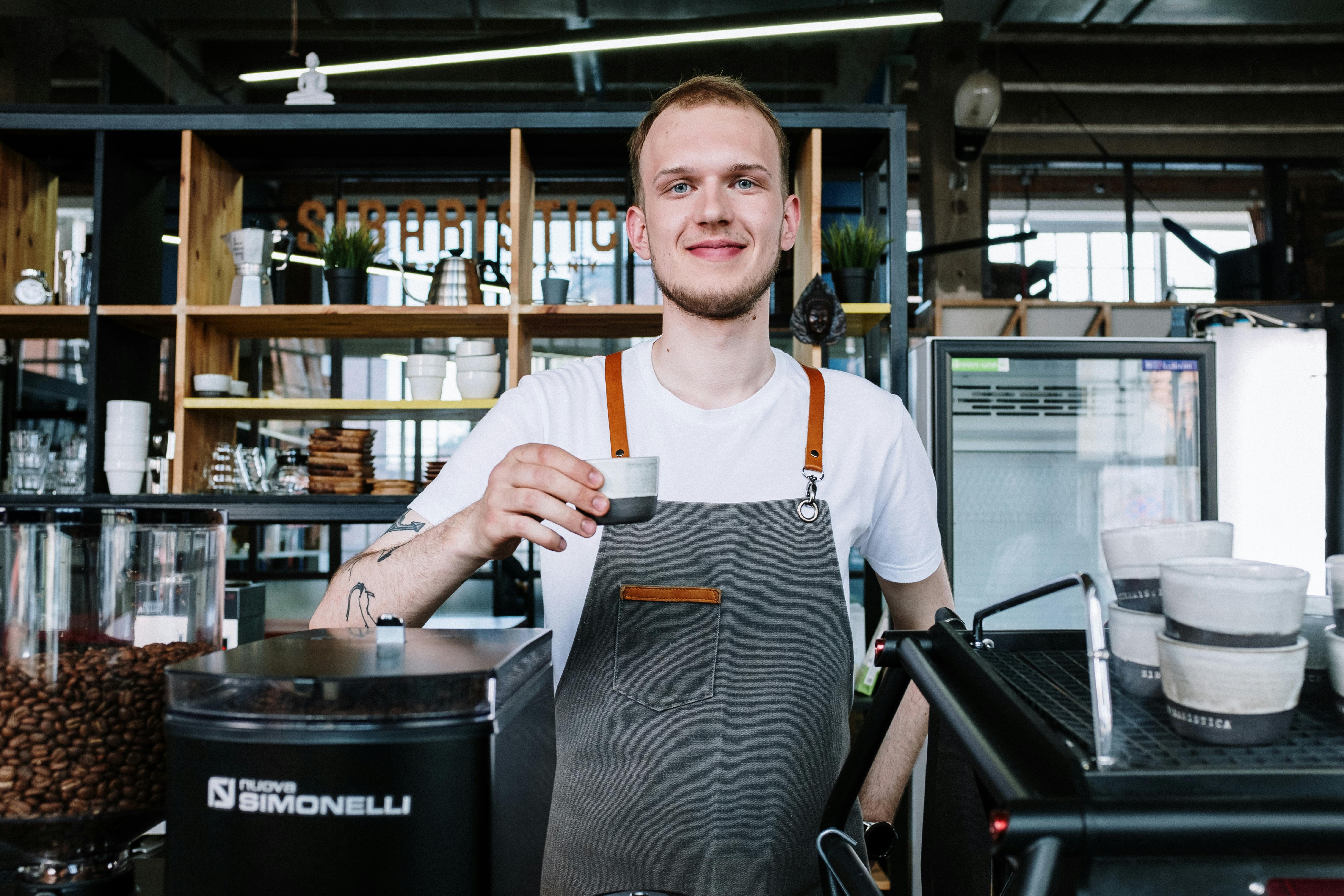 pexels-photo-4790061-4790061 Barista in apron smiling and serving coffee at a modern café. Interior shot with coffee equipment.