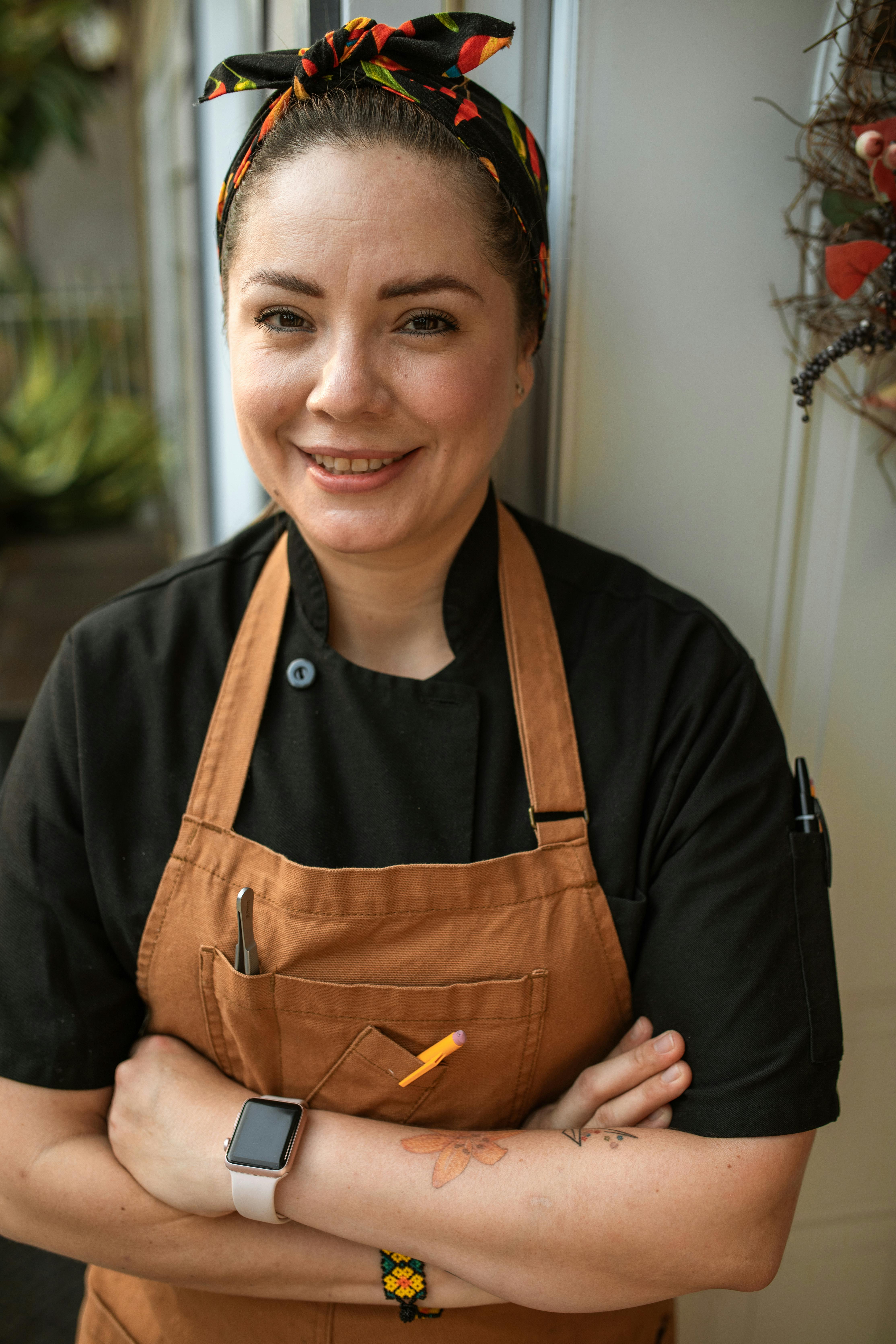 pexels-photo-5737830-5737830 Smiling female chef wearing a brown apron and headband, standing with crossed arms.